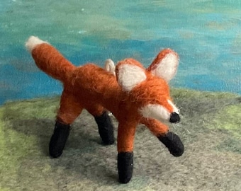May include: A hand-felted fox figurine with a red-orange body, white accents on the face and tail, and black felt feet. The fox is standing on a gray and green surface.
