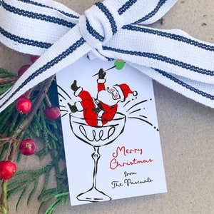 Puede incluir: Una etiqueta de regalo blanca con una ilustraci&oacute;n roja y blanca de Santa Claus cayendo en una copa de champ&aacute;n. El texto en la etiqueta dice "Feliz Navidad de parte de los Pascuale".