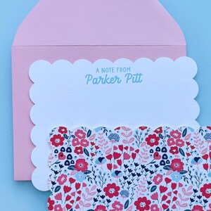 May include: A pink envelope with a scalloped edge holds a matching floral stationery set. The stationery features a white scalloped edge card with the text "A NOTE FROM Parker Pitt" in light blue. The floral pattern includes red, blue, and white flowers.