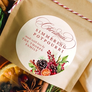 May include: A brown paper bag with a clear window, filled with dried fruit, spices, and greenery. A round sticker reads "Christmas Simmering Potpourri" with botanical illustrations. The bag is tied with red and white twine.