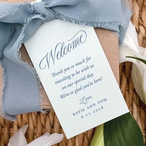 May include: A light blue ribbon tied around a small package with a welcome tag. The tag has the word "Welcome" in elegant script, along with a message and the names Katie and Tom, and the date 6.15.2028.