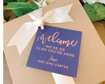 May include: A brown gift box with a white ribbon and a blue tag that says "Welcome, We're so glad you're here, love, Amy and Carter".