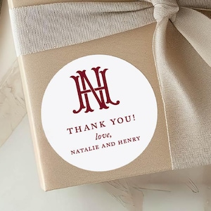 May include: A beige gift box tied with a light brown ribbon and bow. A white circular sticker with burgundy text and a monogram is affixed to the box. The sticker reads "THANK YOU! love, NATALIE AND HENRY."