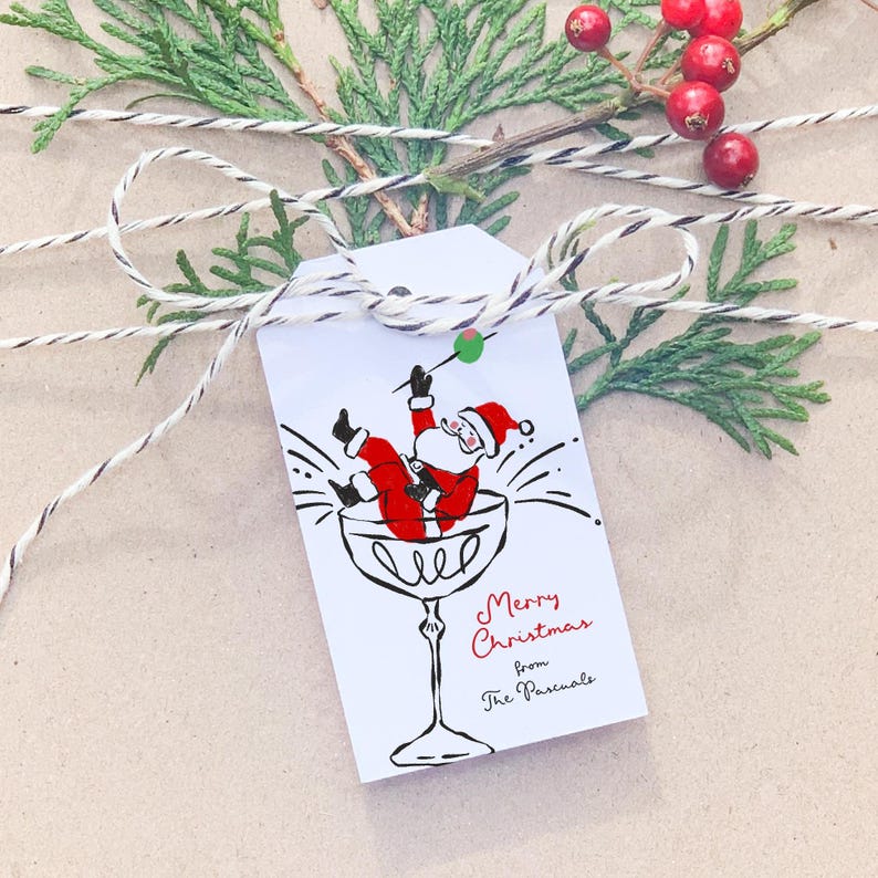 Puede incluir: Una etiqueta de regalo blanca con una ilustraci&oacute;n en negro y rojo de Santa Claus cayendo en una copa de martini. El texto en la etiqueta dice "Feliz Navidad de parte de los Pascuals".