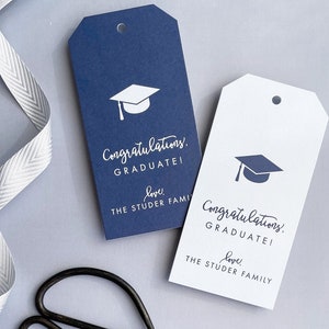 May include: Two graduation gift tags, one navy blue and one white, with a graduation cap graphic and the text "Congratulations! Graduate! love, The Studer Family." A white ribbon is in the background.
