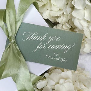 May include: A sage green gift tag with the words "Thank you for coming!" in elegant white script, tied with a light green satin ribbon. The tag is attached to a white envelope, with white flowers in the background.