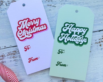 May include: Two gift tags with festive holiday greetings. The tag on the left is white with red and pink lettering that says "Merry Christmas". The tag on the right is light green with dark green lettering that says "Happy Holidays". Both tags have blank spaces for "To:" and "From:".