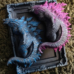 Puede incluir: Escultura enmarcada con dos figuras estilizadas de dragón. Una es azul, la otra gris oscuro con detalles en rosa. El marco es gris oscuro con detalles ornamentales, sobre un fondo texturizado.