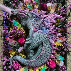 May include: A framed artwork featuring a sculpted dragon with a textured, grey body and purple and pink accents. The dragon is surrounded by colourful crystals, dried flowers, and greenery, all set within a black frame.