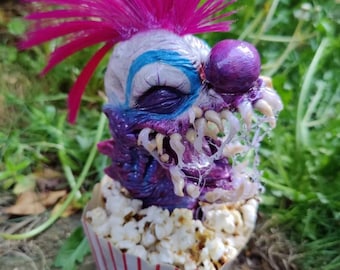 May include: A sculpted clown figure with a bright pink mohawk, purple skin, and a large purple nose, emerging from a white and red striped popcorn container filled with popcorn. The figure features sharp, translucent teeth and blue eye makeup.