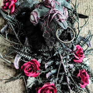 May include: A black and silver animal skull with a ring attached to the nose, surrounded by dark green foliage and red roses.