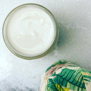 May include: A white cream in a clear glass jar with a wide mouth. The jar is sitting on a white marble surface.
