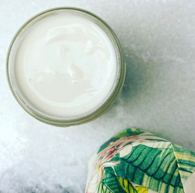 May include: A white cream in a clear glass jar with a wide mouth. The jar is sitting on a white marble surface.