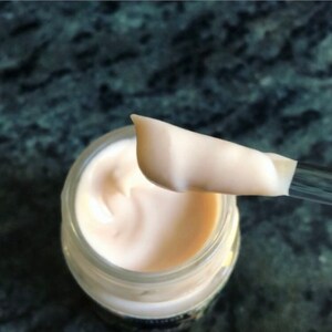 May include: A close-up of a white cream in a glass jar. A spatula is scooping out the cream, showing its smooth, creamy texture.