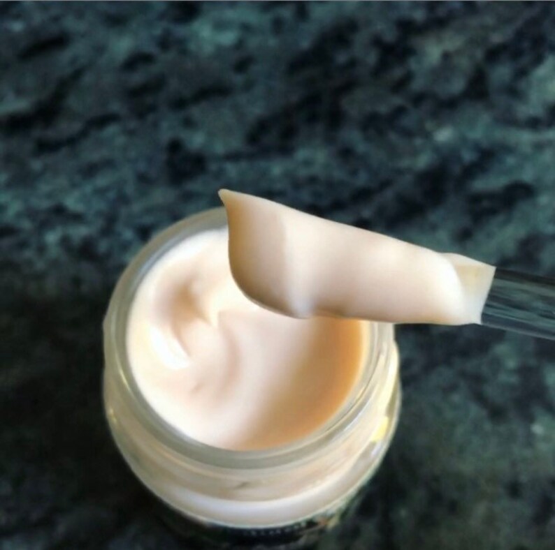 May include: A close-up of a white cream in a glass jar. A spatula is scooping out the cream, showing its smooth, creamy texture.