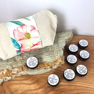 May include: A set of six small, round, black tins containing perfume cologne samples. The tins are arranged on a wooden surface with a beige canvas bag with a floral design in the background. The tins are labelled with the names of the scents: Anahata Heart, Amber, Cardamom Rose, Sandalwood Forest, Patchouli Frangipani, and Orange Blossom.