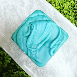 May include: A square-shaped bar of soap with a swirling, marbled pattern in shades of teal and blue. The soap has a speckled, glittery appearance.