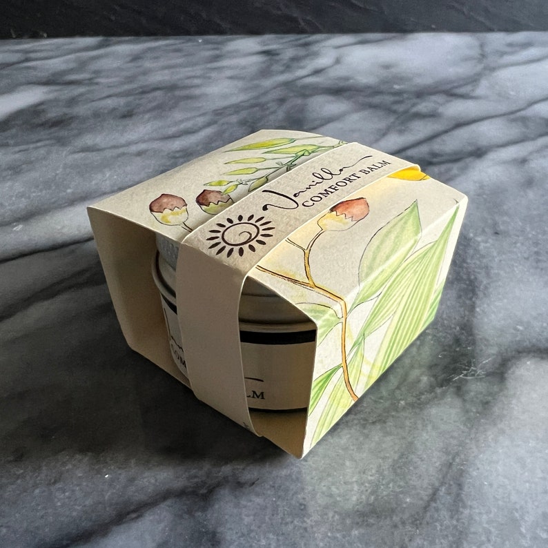 May include: A small, white tin with a black lid containing a vanilla comfort balm. The tin is packaged in a cream-colored box with a floral design and the text "Vanilla Comfort Balm".