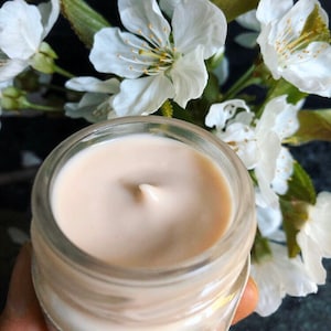 May include: A close-up of a glass jar filled with a light beige cream. The cream has a smooth, swirl-like texture. The jar is partially obscured by white cherry blossoms.
