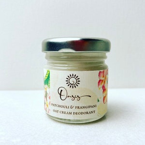 May include: A small glass jar with a silver lid containing a white cream deodorant. The label is white with a floral design and the text "Oasis Patchouli & Frangipani Oat Cream Deodorant".