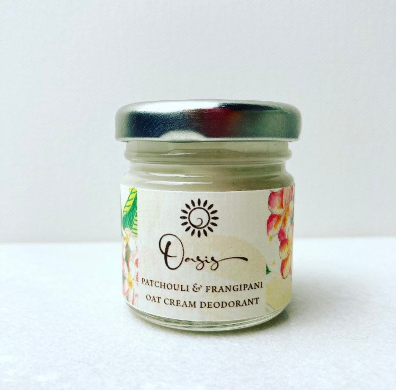 May include: A small glass jar with a silver lid containing a white cream deodorant. The label is white with a floral design and the text "Oasis Patchouli & Frangipani Oat Cream Deodorant".