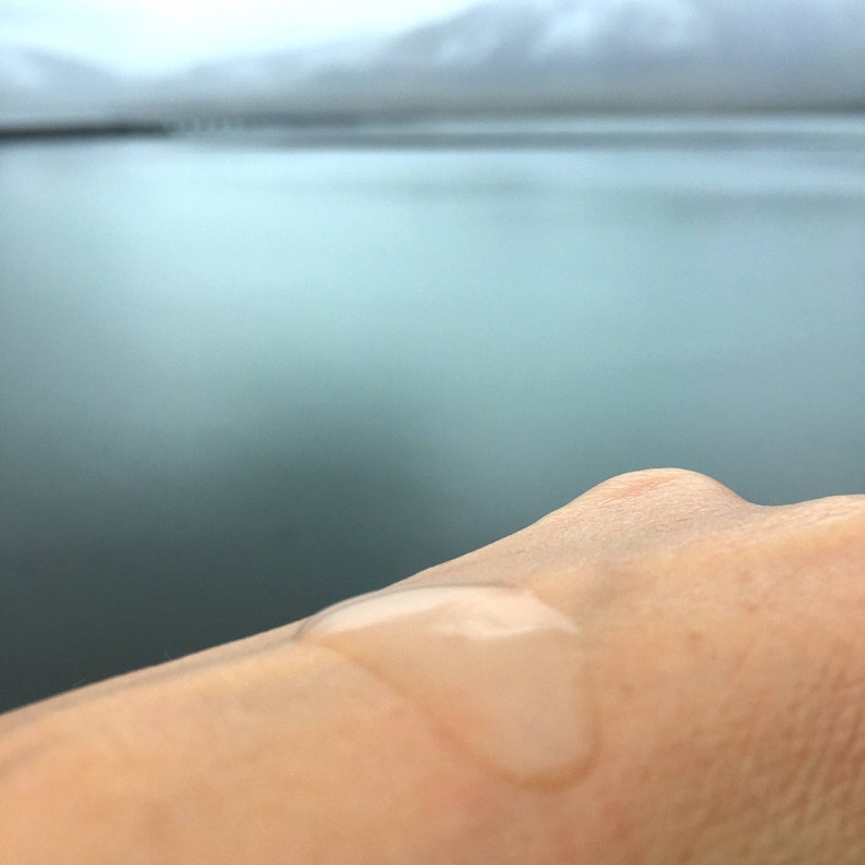 May include: A close-up of a hand with a drop of water on it. The background is a blurry image of a body of water and mountains.