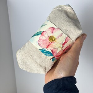 Pu&ograve; includere: Una pochette beige in tela con cerniera e un design floreale. Il design presenta fiori rosa e gialli con foglie verdi.