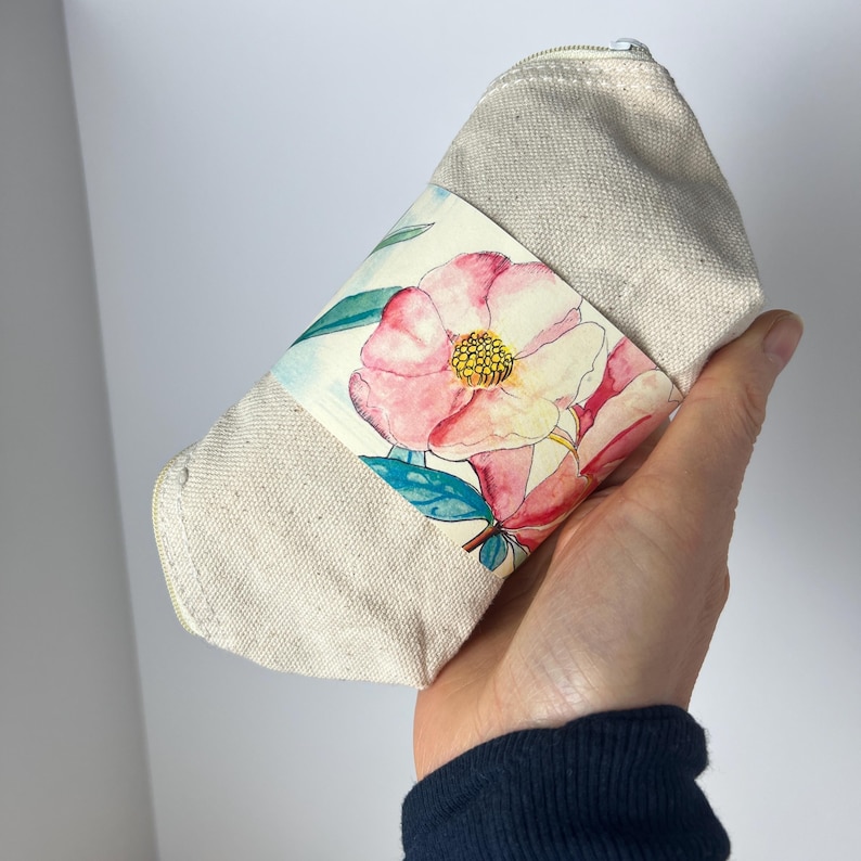 Pu&ograve; includere: Una pochette beige in tela con cerniera e un design floreale. Il design presenta fiori rosa e gialli con foglie verdi.