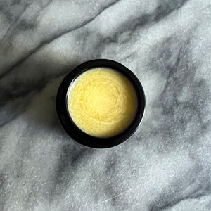 May include: A small black jar with a lid containing a yellow balm or ointment. The balm has a swirl pattern.