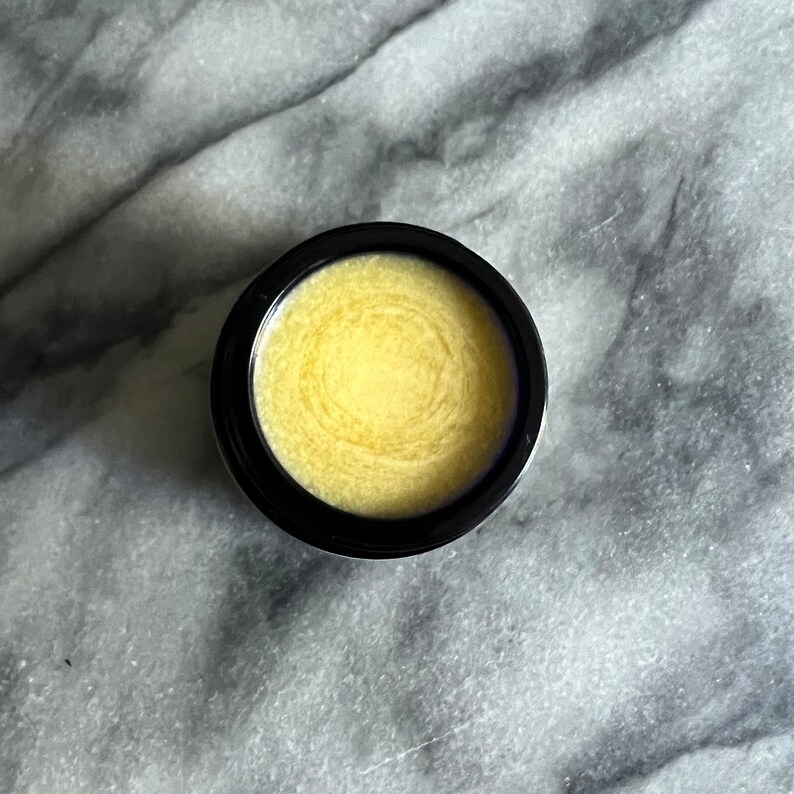 May include: A small black jar with a lid containing a yellow balm or ointment. The balm has a swirl pattern.