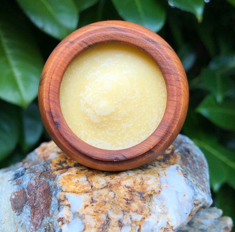 May include: A small wooden bowl filled with a pale yellow, creamy substance. The bowl is sitting on a rock with a green leafy background.