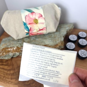 Pu&ograve; includere: Una piccola borsa di tela bianca con un motivo floreale. La borsa &egrave; aperta e si vede una piccola etichetta di carta bianca. L'etichetta riporta il testo "Anahata Heart Solid Perfume & Colorus" e elenca gli ingredienti. La borsa &egrave; appoggiata su una superficie di legno.