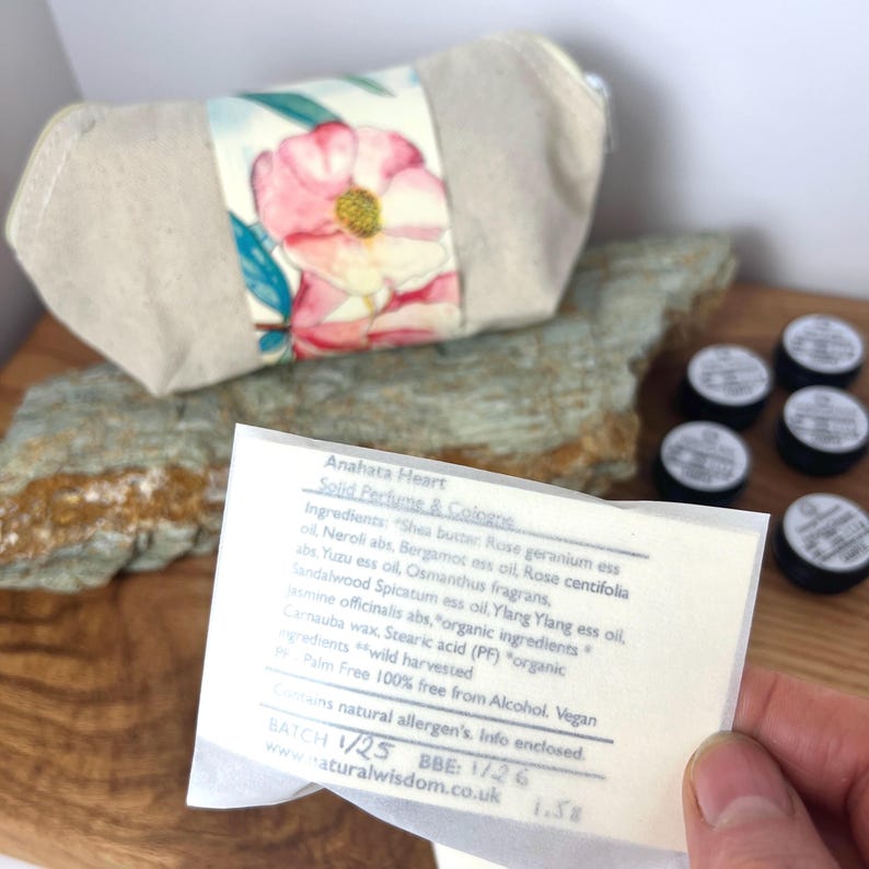 Pu&ograve; includere: Una piccola borsa di tela bianca con un motivo floreale. La borsa &egrave; aperta e si vede una piccola etichetta di carta bianca. L'etichetta riporta il testo "Anahata Heart Solid Perfume & Colorus" e elenca gli ingredienti. La borsa &egrave; appoggiata su una superficie di legno.