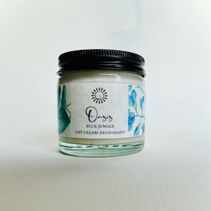 May include: A glass jar of Oasis Blue Jungle Oat Cream Deodorant. The jar has a black lid and a white label with blue floral illustrations. The label reads "Oasis Blue Jungle Oat Cream Deodorant".