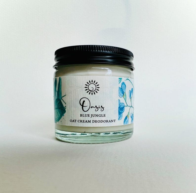 May include: A glass jar of Oasis Blue Jungle Oat Cream Deodorant. The jar has a black lid and a white label with blue floral illustrations. The label reads "Oasis Blue Jungle Oat Cream Deodorant".