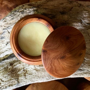 May include: A wooden container with a lid, containing a light yellow balm. The container has a natural wood grain pattern.