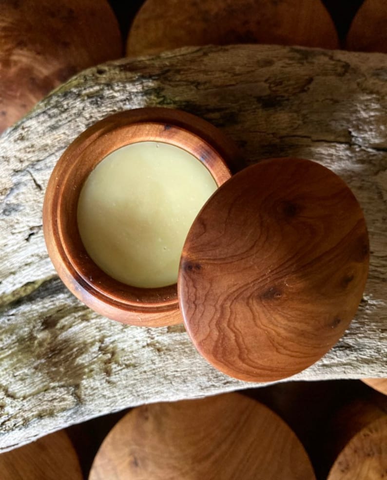 May include: A wooden container with a lid, containing a light yellow balm. The container has a natural wood grain pattern.