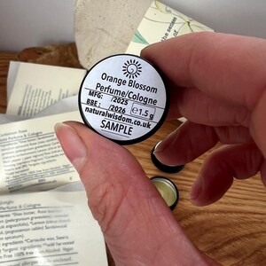 Pu&ograve; includere: Un piccolo contenitore rotondo nero con un'etichetta bianca che recita "Orange Blossom Perfume/Cologne" con il testo "MFG: /2025 BBE: /2026 e1.5 g naturalwisdom.co.uk SAMPLE".
