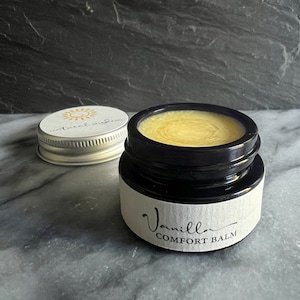 May include: A small glass jar with a black lid and a white label that reads "Vanilla Comfort Balm". The jar is filled with a yellow balm.