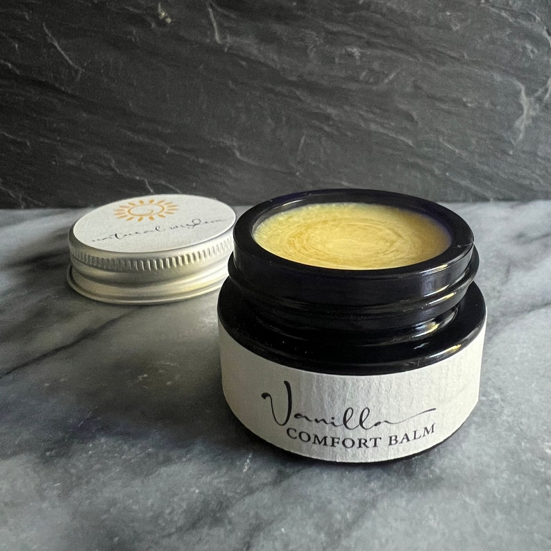 May include: A small glass jar with a black lid and a white label that reads "Vanilla Comfort Balm". The jar is filled with a yellow balm.