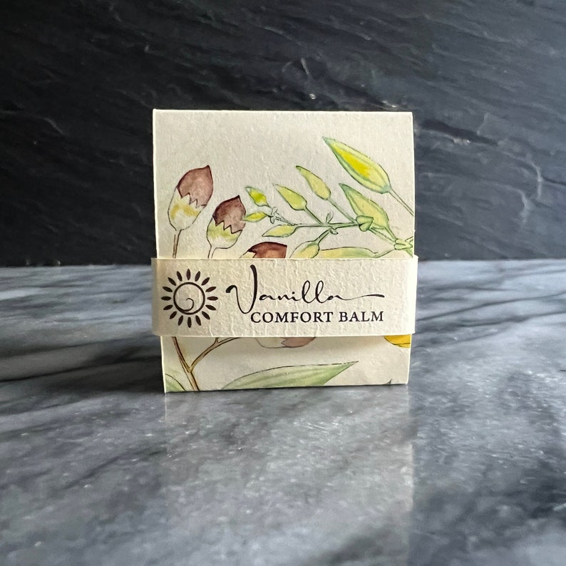 May include: A small, rectangular box with a white background and a floral design. The box has a label that says "Vanilla Comfort Balm".