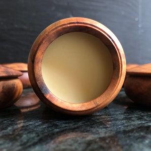Könnte beinhalten: Ein Holzbehälter mit Deckel, gefüllt mit einem hellbeigen Balsam. Der Behälter hat eine dunkelbraune Holzmaserung und eine glatte, polierte Oberfläche.