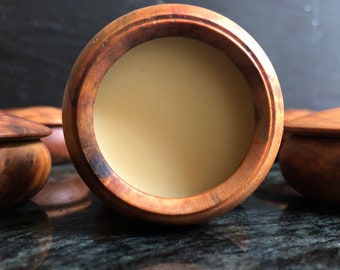 May include: A wooden container with a lid, filled with a light-colored balm or cream. The container has a natural wood grain and a smooth, rounded shape.