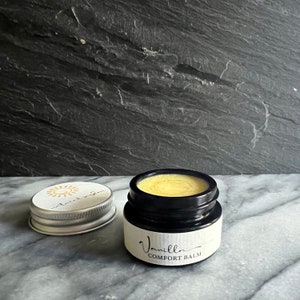 May include: A small glass jar with a black lid and a white label that reads "Vanilla Comfort Balm". The jar is filled with a yellow balm.