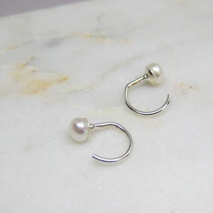 Puede incluir: Un par de pendientes de aro pequeños, de color plateado, con una sola perla blanca redonda en la parte superior de cada pendiente. Los pendientes se muestran sobre una superficie de mármol blanco y gris.