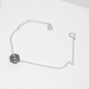 May include: A silver chain bracelet featuring a dark gray hexagonal nut charm. The bracelet has a delicate chain design with a lobster clasp closure and an adjustable chain. The bracelet is on a white background.