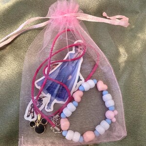 May include: A pink organza bag with a pink ribbon tie contains a beaded bracelet with pink, blue, and white beads. The bracelet has a heart charm and a Mickey Mouse charm. A sticker of a Disney princess is also included.