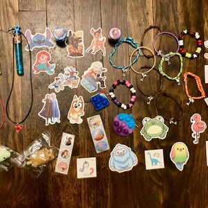 May include: A collection of colorful bracelets, necklaces, and stickers featuring Disney characters like Stitch, Mickey Mouse, and a unicorn. There are also a few small toys, including a frog, a flamingo, and a unicorn.