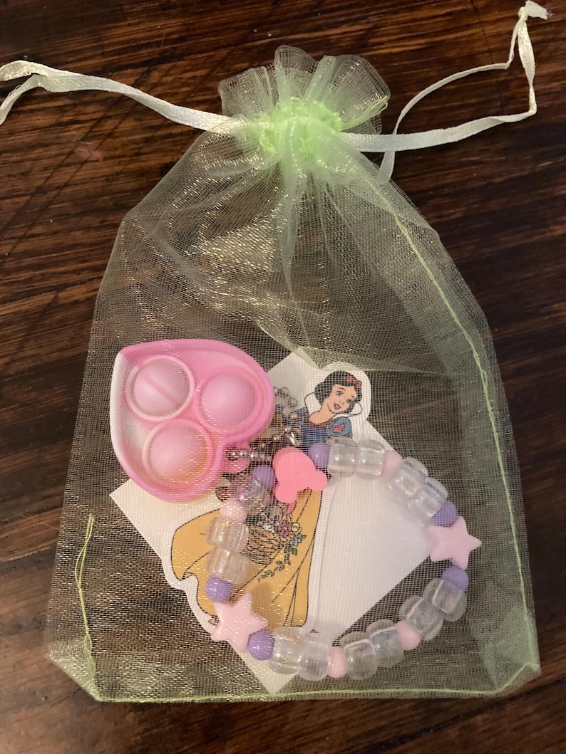 May include: A green mesh bag with a pink heart-shaped pop-it toy, a pink and purple beaded bracelet with star charms, and a sticker featuring Snow White from Disney.