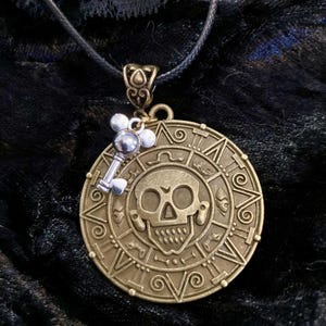 May include: A gold-toned necklace with a round pendant featuring a skull and a key charm. The pendant has intricate details and a dark background.